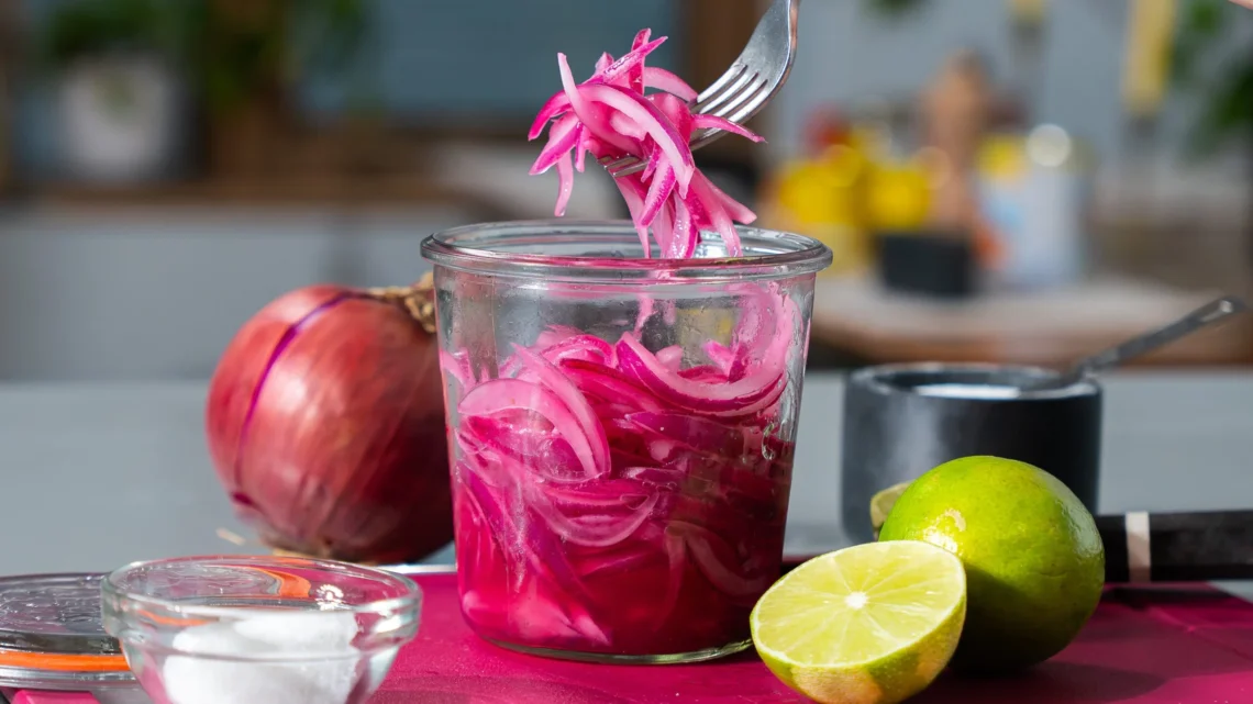 jar of bright pink pickled red onions with a fork lifting onion slices, lime and red onion nearby