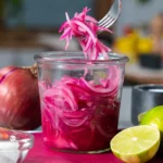 jar of bright pink pickled red onions with a fork lifting onion slices, lime and red onion nearby