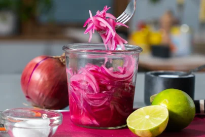 jar of bright pink pickled red onions with a fork lifting onion slices, lime and red onion nearby