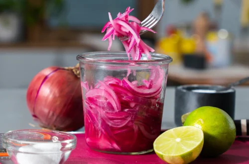 jar of bright pink pickled red onions with a fork lifting onion slices, lime and red onion nearby