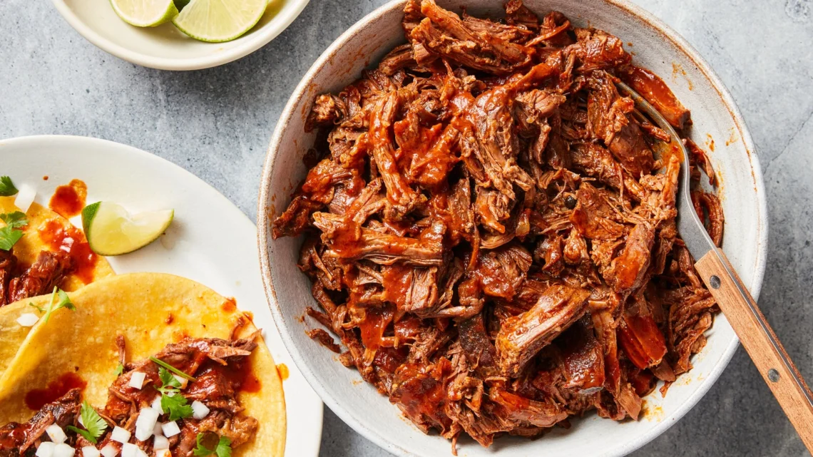 shredded beef barbacoa in a bowl with lime wedges and tacos on the side