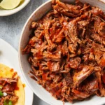 shredded beef barbacoa in a bowl with lime wedges and tacos on the side