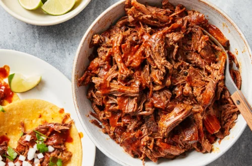 shredded beef barbacoa in a bowl with lime wedges and tacos on the side