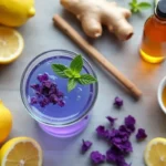 purple wellness drink in a glass with mint, surrounded by lemon, ginger, cinnamon stick, honey, and cucumber slices