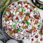 muddy buddy puppy chow mix in a bowl with powdered sugar, pretzels, mini marshmallows and red green candy