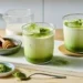 two glasses of iced matcha latte with milk, matcha powder, honey dipper and whisk on a tray