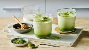 two glasses of iced matcha latte with milk, matcha powder, honey dipper and whisk on a tray